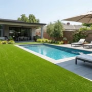 Artificial turf surrounds a modern pool area with lounge chairs and umbrella.